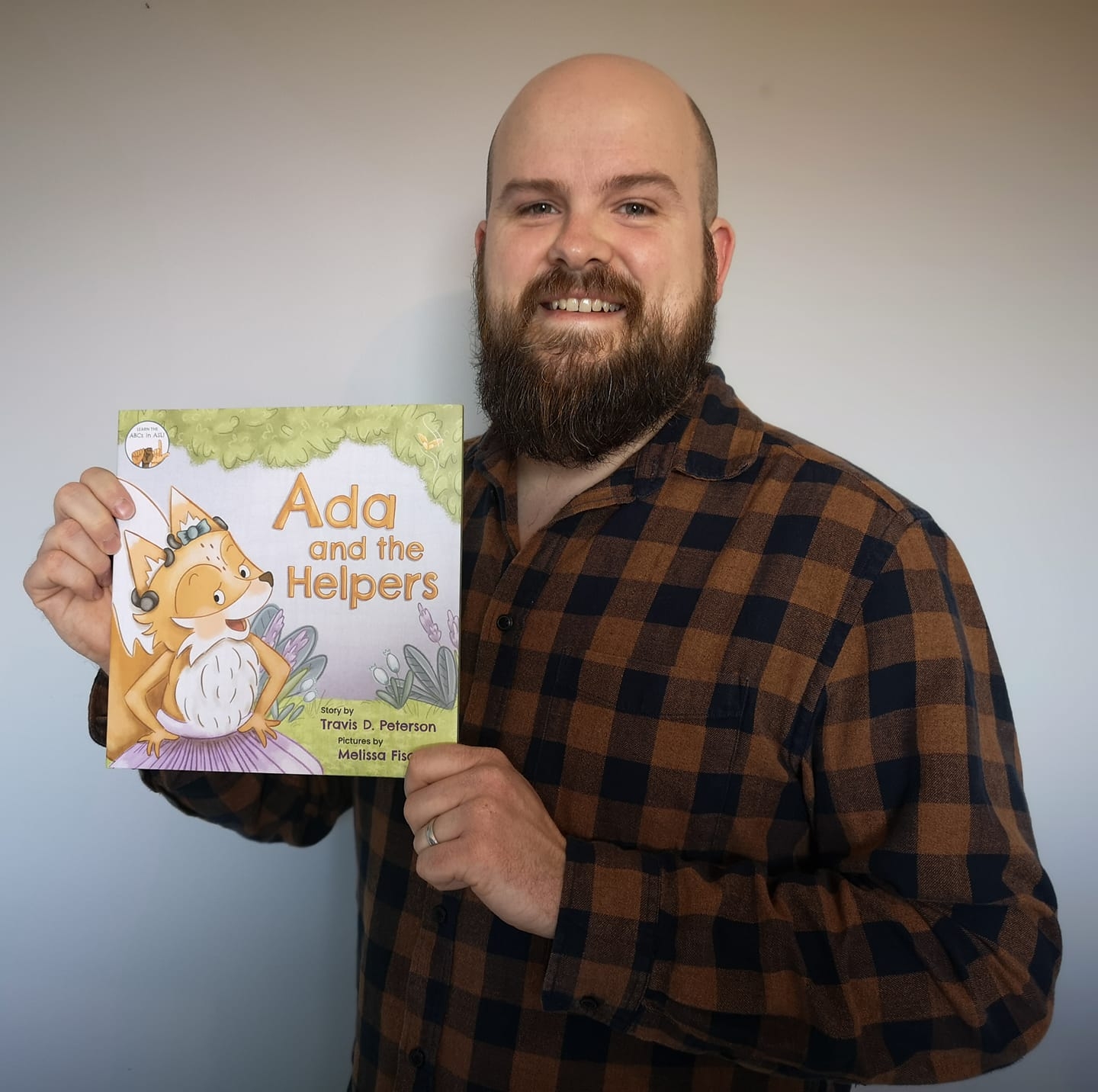 Author Travis D. Peterson wearing a flannel shirt and holding a copy of Ada and the Helpers children's book